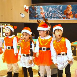 クリスマスの飾り付けをバックに、サンタの衣装を着て一生懸命にお歌を歌っている子どもたちの発表会の様子。