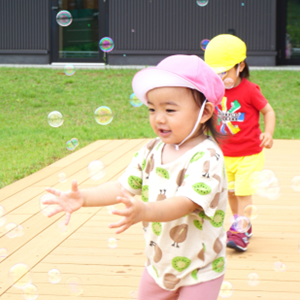 ウッドデッキの上で、ふわふわと飛んでくるたくさんのシャボン玉を、両手を広げて捕まえようとしている女の子。